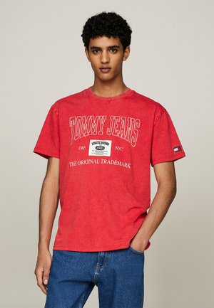 Camiseta de algodón roja con mangas cortas, que presenta el texto blanco "TOMMY JEANS" y un gráfico de logo. Combinada con unos vaqueros de denim azul.