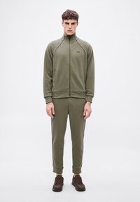 Φόρμα σε ελιά πράσινο χρώμα με φερμουάρ, με αντίθετες μαύρες ρίγες, μανσέτες και τελείωμα με ριμπ ύφανση, ζευγά�ρι καφέ αθλητικά παπούτσια, απλός σχεδιασμός, χωρίς ορατά λογότυπα.