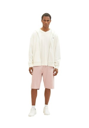 Haut à fermeture éclair blanc sur un t-shirt blanc, associé à un short rose clair et des baskets blanches. Design minimaliste, coupe décontractée.