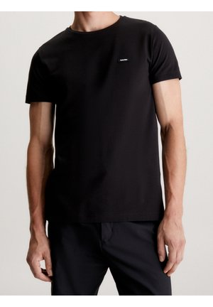 Männliches Modell trägt ein schlichtes schwarzes T-Shirt mit kurzen Ärmeln und kleinem Calvin Klein-Logo auf der Brust, kombiniert mit schwarzen Hosen vor neutralem Hintergrund.