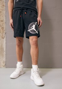 Shorts de sport noirs avec un logo Jordan blanc et un design circulaire, dotés d'une taille élastique et de cordons orange. Portés avec des baskets blanches.