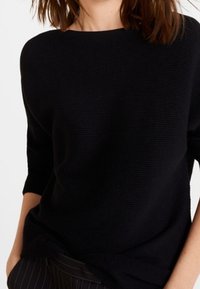 Pull en maille noire texturée avec un large col bateau et des manches trois-quarts. Présente une coupe décontractée, sans motifs ni accents visibles.