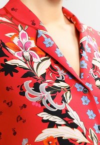 Red floral blouse with short collar, featuring large pink and blue flowers, green leaves, and black accents. Smooth, textured fabric.