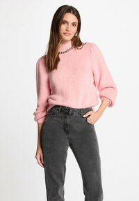 Lichtroze fuzzy sweater met een hoge hals en ingezette mouwen, gecombineerd met donkergrijze high-waisted jeans. Heeft een zilveren kraal ketting.