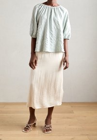 Blouse texturée bleu clair avec des manches bouffantes, associée à une jupe fluide de couleur crème. Les chaussures sont des sandales ouvertes beiges avec des brides croisées.