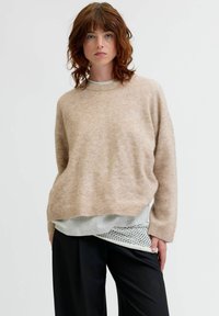 Beżowy sweter dzianinowy o luźnym kroju, z prążkowanym dekoltem i mankietami, noszony na szarym T-shircie z teksturowanym brzegiem, w zestawieniu z czarnymi spodniami.