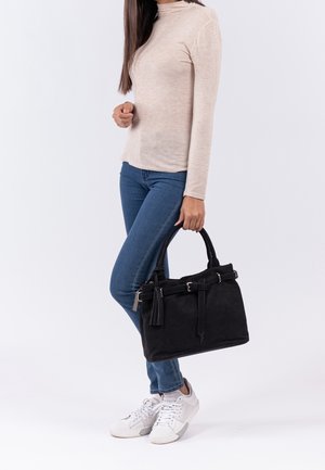 Femme portant un haut à manches longues beige, un jean bleu, des baskets blanches, tenant un sac à main noir orné de pampilles sur un fond blanc uni.