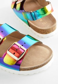 Regnbågsfärgade sandaler har en glänsande metallisk ovandel och två justerbara remmar med silverfärgade spännen. Sulan är vit och strukturerad.