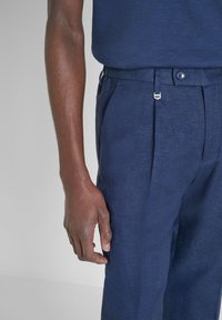 Marineblauwe broek van textuurstof, met voorplooien, een zilveren accent op de tailleband en een knoopsluiting.