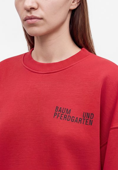 Femme portant un sweat-shirt rouge avec un texte noir « "BAUM UND PFERDGARTEN" » sur la poitrine, sur un fond blanc.