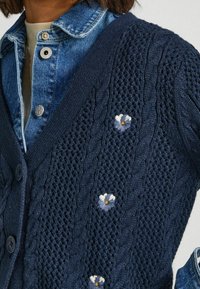 Navy knitted cardigan with cable and textured patterns, featuring embroidered blue and white flowers. Buttoned front and V-neck design.