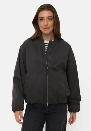 Femme portant une veste bomber noire zippée par-dessus une chemise rayée et un pantalon noir, debout devant un fond clair uni.