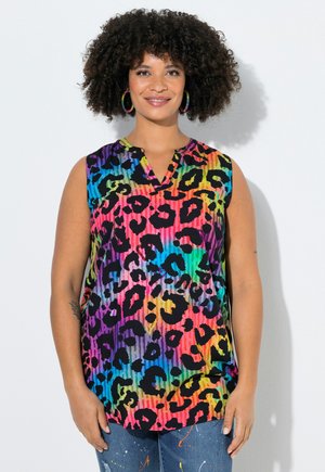 Kvinde med krøllet hår iført ærmeløs regnbueleopardprint top og farverige store øreringe, stående mod hvid baggrund.