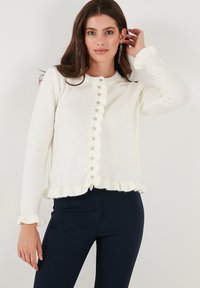 Weißer gestrickter Cardigan mit einer Rüsche am Saum und an den Bündchen, mit runden Knöpfen in der Mitte. Weiches Material, schlanke Passform und lange Ärmel.