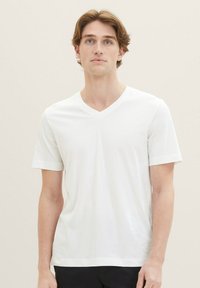 Weißes V-Ausschnitt T-Shirt aus weichem Stoff mit kurzen Ärmeln und subtiler Textur. Minimalistisches Design mit kleinem Logo nahe dem Saum.
