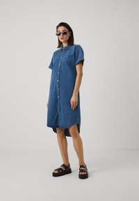 Φόρεμα πουκαμίσα από τζιν σε μεσαία μπλε απόχρωση, με κοντά μανίκια, κουμπιά μπροστά και μήκος μέσης με ελαφρώς καμπυλωτή πλάτη. Συνδυασμένο με σκούρες σανδάλια.
