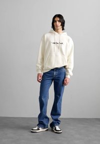 Creme hoodie med frontlomme og sort logo, parret med blå jeans og sort-hvide sneakers. Enkelt gråt baggrund.