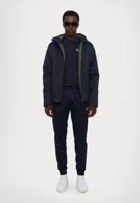 Completo sportivo della marina con giacca con zipper e fodera verde, polo, pantaloni con vita elastica e sneakers bianche.