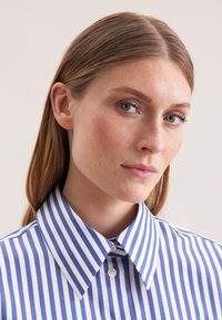 Jonge vrouw met steil lichtbruin haar, gekleed in een blauw-wit verticaal gestreept overhemd, kijkt iets opzij.