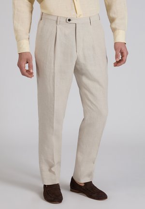 Mann trägt beige Faltenhose, hellgelbes Hemd und dunkelbraune Wildleder-Loafer, steht vor einem schlichten hellgrauen Hintergrund.