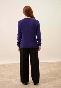 Neulepusero purppurasta langasta, jossa on tekstuuri, pyöreä kaula-aukko ja pitkät hihat, yhdistettynä laajoihin mustiin housuihin.