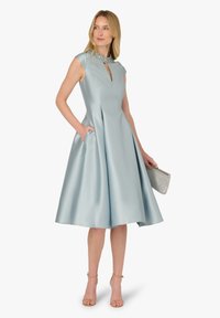 Jasnoniebieska sukienka z satyny z dopasowanym górnym stanem, krótkimi rękawami, dekoltem z wycięciem na klucz, plisowanym dołem i kieszeniami po bokach. Biżuteria w srebrnym odcieniu i kopertówka.