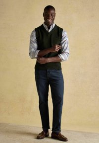 Usmiaty muž v tmavozelenom bezrukávovom svetri cez kostkovanú košeľu, tmavých džínsach a hnedých mokasínach stojí so založenými rukami proti béžovej stene.