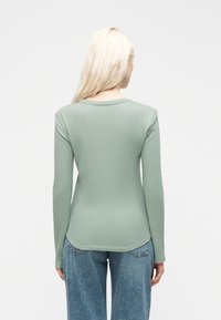 Γυναίκα με μακριά ξανθά μαλλιά φοράει μπλούζα μέντας με μακριά μανίκια και μπλε τζιν, στέκεται γυρισμένη με την πλάτη σε λευκό φόντο.