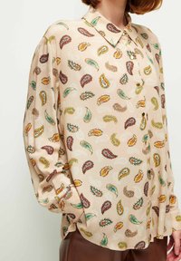 Camisa beige claro con un patrón de paisley en rojo, verde y amarillo, con un collar puntiagudo y cierre de botones. Tejido suave y texturizado.