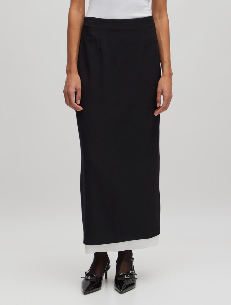 Zwarte zwijgende rok van vloerlengte, gemaakt van gladde stof met een aansluitende tailleband. Beschikt over een contrasterende witte zoom. Gemaakt met puntige zwarte schoenen.