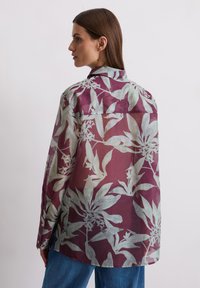 Long-sleeve shirt made of sheer fabric, featuring a purple background with light green floral patterns. Button details on sleeves.