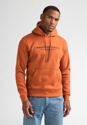 Homme portant un sweat à capuche orange Petrol Industries et un jean bleu, debout avec une expression neutre face à un fond clair uni.