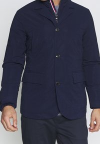 Giacca leggera di colore blu navy, con tessuto testurizzato, tasche frontali diagonali, chiusura a bottoni e colletto sagomato.