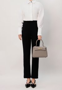 Borsa strutturata grigia con hardware dorato, tenuta con un manico curvo. Il modello indossa una blusa bianca con colletto e pantaloni neri aderenti, con scarpe nere.