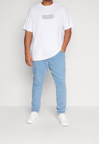 Camiseta blanca de algodón con el logo gris de "Calvin Klein", combinada con unos vaqueros de mezclilla azul claro. El modelo lleva zapatillas blancas. Estilo casual, ajuste relajado.