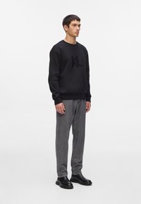 Sudadera negra con logo texturizado, puños y dobladillo acanalados. Combinada con pantalones grises y zapatos negros, de pie contra un fondo neutral.