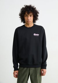 Felpa nera con polsini e orlo a coste, con il logo rosa "Dickies" sul lato sinistro del petto. Modello abbinato a pantaloni verde oliva.