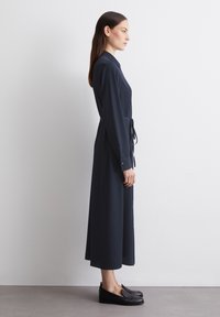 Abito lungo a maniche con colletto, di colore blu navy, con vita contrassegnata da una cintura e tasche laterali. Tessuto morbido; abbinato a scarpe basse nere.