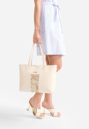 Donna in abito azzurro chiaro che porta una borsa tote crema con stampa di mappa, indossa sandali crema con tacco e cinturini con fibbia.