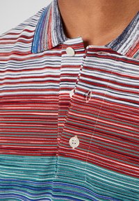 Gros plan d’un polo rayé aux lignes horizontales blanches, rouges, bleues et turquoises avec des boutons blancs sur la patte de boutonnage et le col.