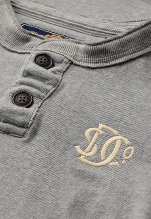 Camisa polo de algodón gris con cuello acanalado, dos botones oscuros y logotipo bordado en color crema. Textura suave con un sutil patrón jaspeado.