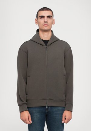 Dark green zip-up hoodie made of soft fabric, featuring a front zipper, large hood, and side pockets, paired with denim jeans.