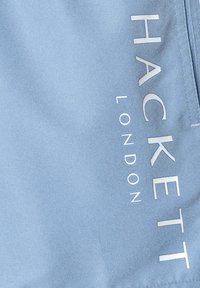 Svetlomodrá látka s textom "HACKETT LONDON" v bielych, vertikálnych písmenách. Textúra vyzerá hladko s jemným leskom.