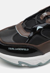 Fekete lakkbőr és hálós felsőrésszel, barna bőr kiegészítőkkel, fehér talppal, és világoskék "KARL LAGERFELD" logóval rendelkező sportcipő.