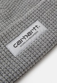 Berretto lavorato a maglia grigio in filato spesso con una texture a costine. Presenta un'etichetta rettangolare bianca con la scritta "carhartt WORK IN PROGRESS."