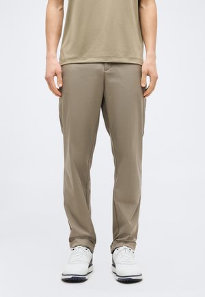 Άνδρας με μπεζ casual παντελόνι και λευκά αθλητικά παπούτσια, στέκεται μπροστά σε ουδέτερο φόντο με τα χέρια χαλαρά στα πλάγια.