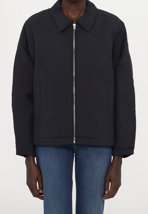 Person iført en sort lynlåstrøje med krave og lange ærmer, kombineret med blå jeans mod en ensfarvet baggrund.
