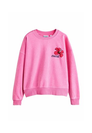 Tânără stă pe o plajă cu nisip aproape de ocean, purtând un hanorac roz cu floare de hibiscus și logo Coca-Cola și pantaloni scurți corali.