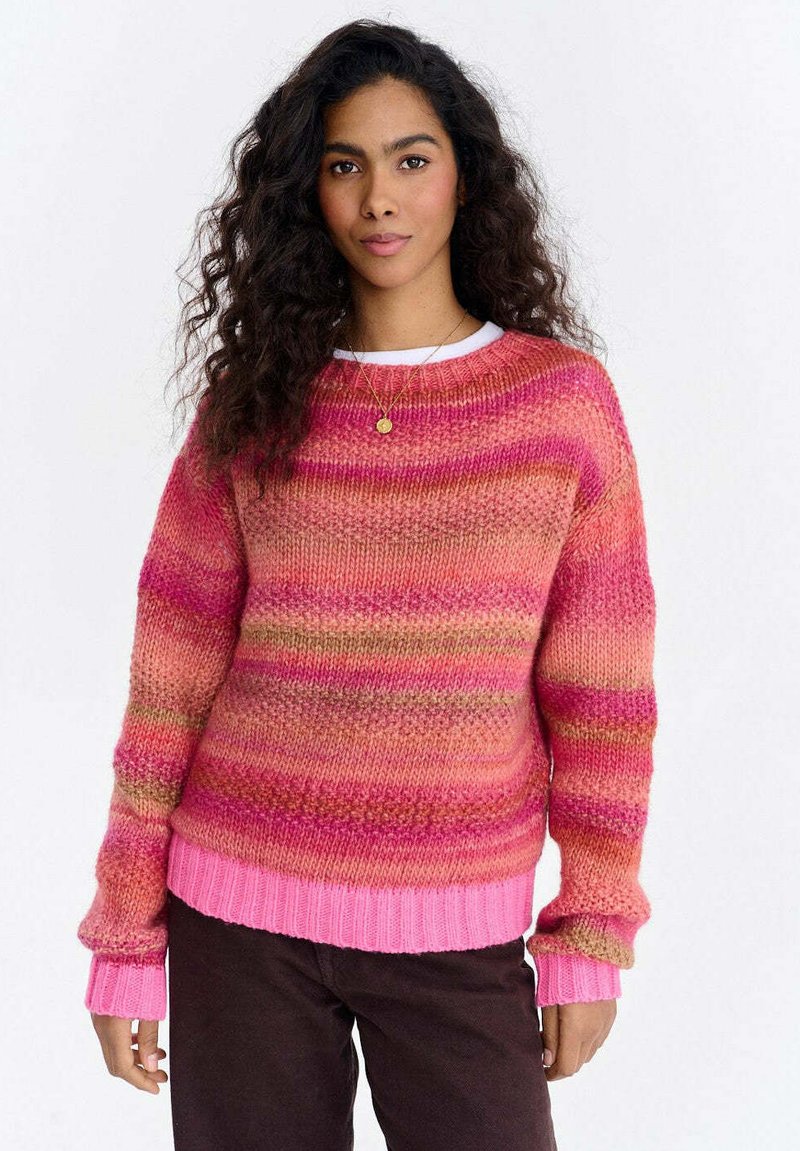 Kobieta z długimi kręconymi włosami, ubrana w wielokolorowy różowo-pomarańczowy sweter na drutach i ciemne spodnie, stoi na tle jednolitego białego tła.