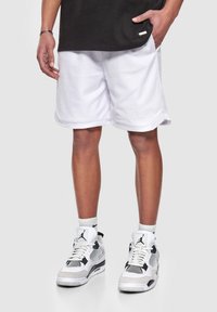 Shorts en maille blanche avec une surface texturée, présentant un ourlet arrondi et un détail de marque subtil. Associés à des baskets de sport noir et blanc.